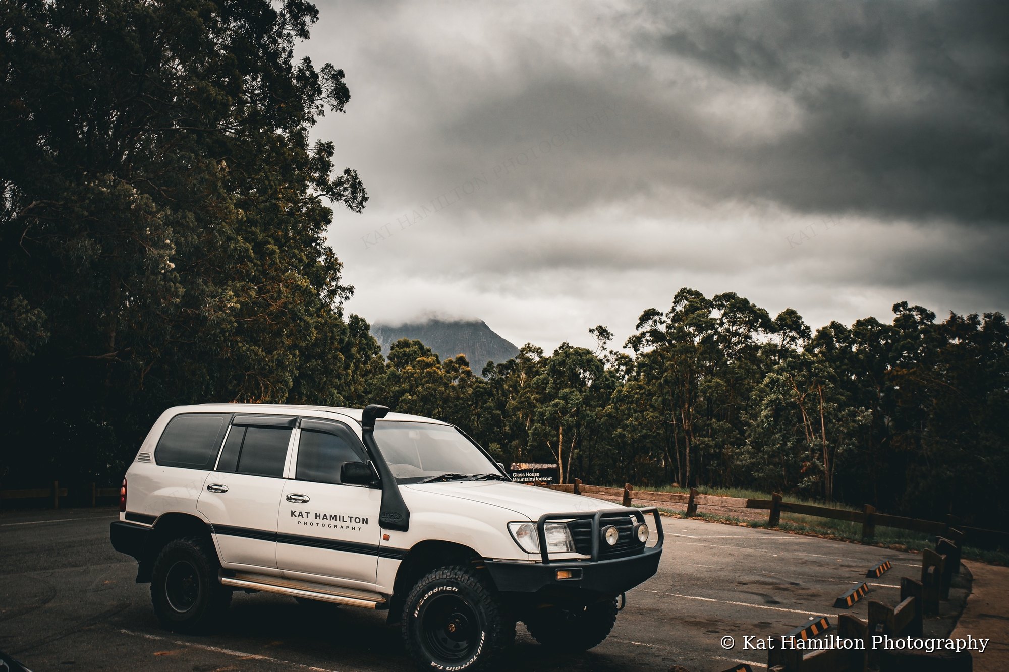A white LandCruiser with Kat Hamilton Photography lettering on the door, parked at a lookout beneath moody clouds, with a mountain peak rising behind the trees.
