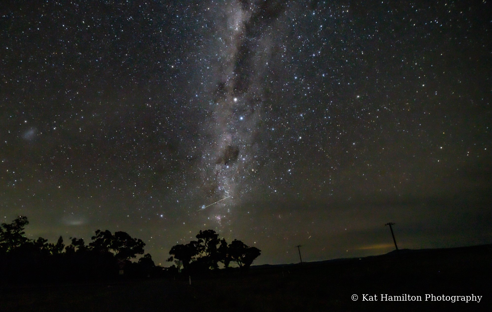 The Milky Way streaking down towards trees on the horizon, with a thin meteor trail beside it and power poles silhouetted on the right.