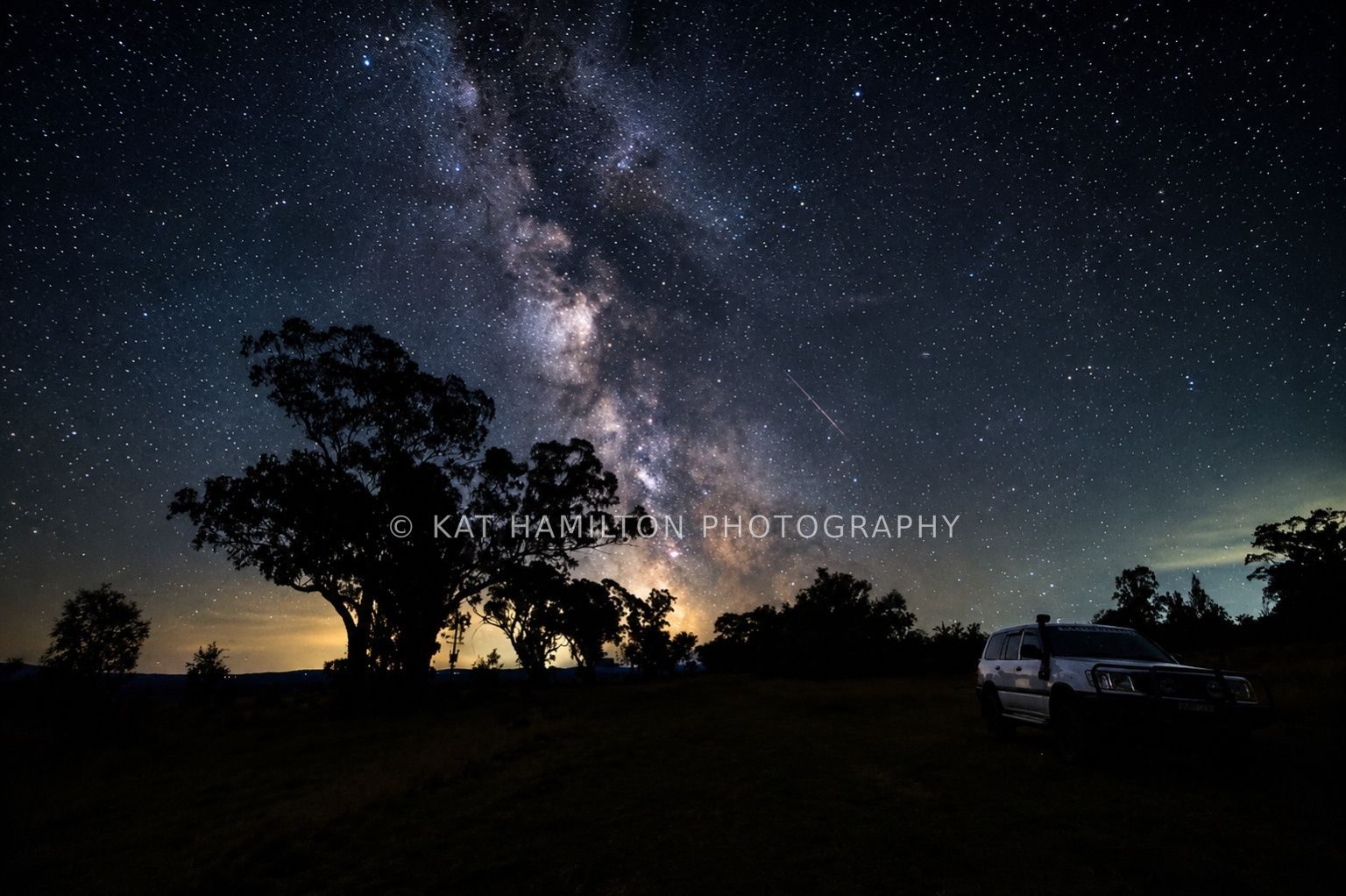 The Milky Way above silhouetted trees and a LandCruiser beneath a star-filled night sky.