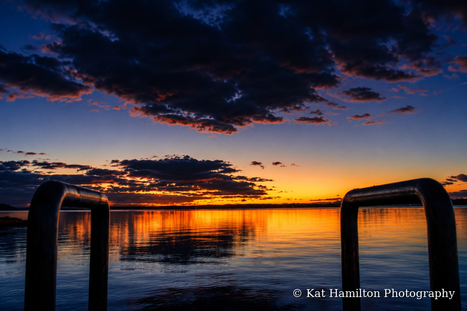 Orange sunset light reflected on calm water under dark clouds, framed by two steel jetty rails.